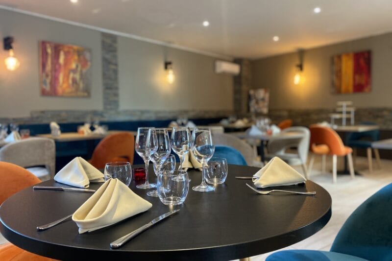 Salle du restaurant dressée en soirée avec tables rondes, verres à vin et chaises colorées