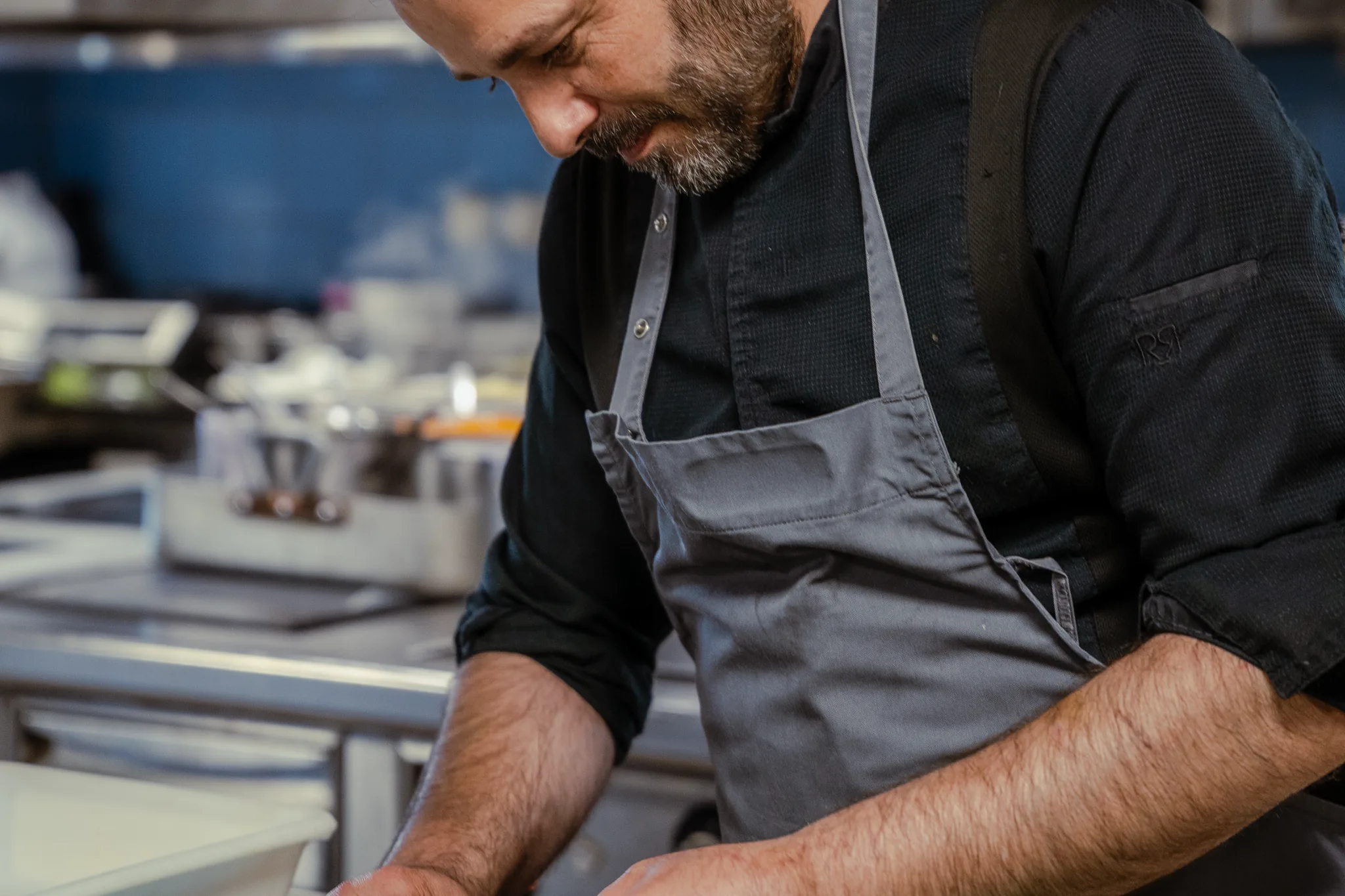Le chef en cuisine, tablier gris, penché sur son plan de travail