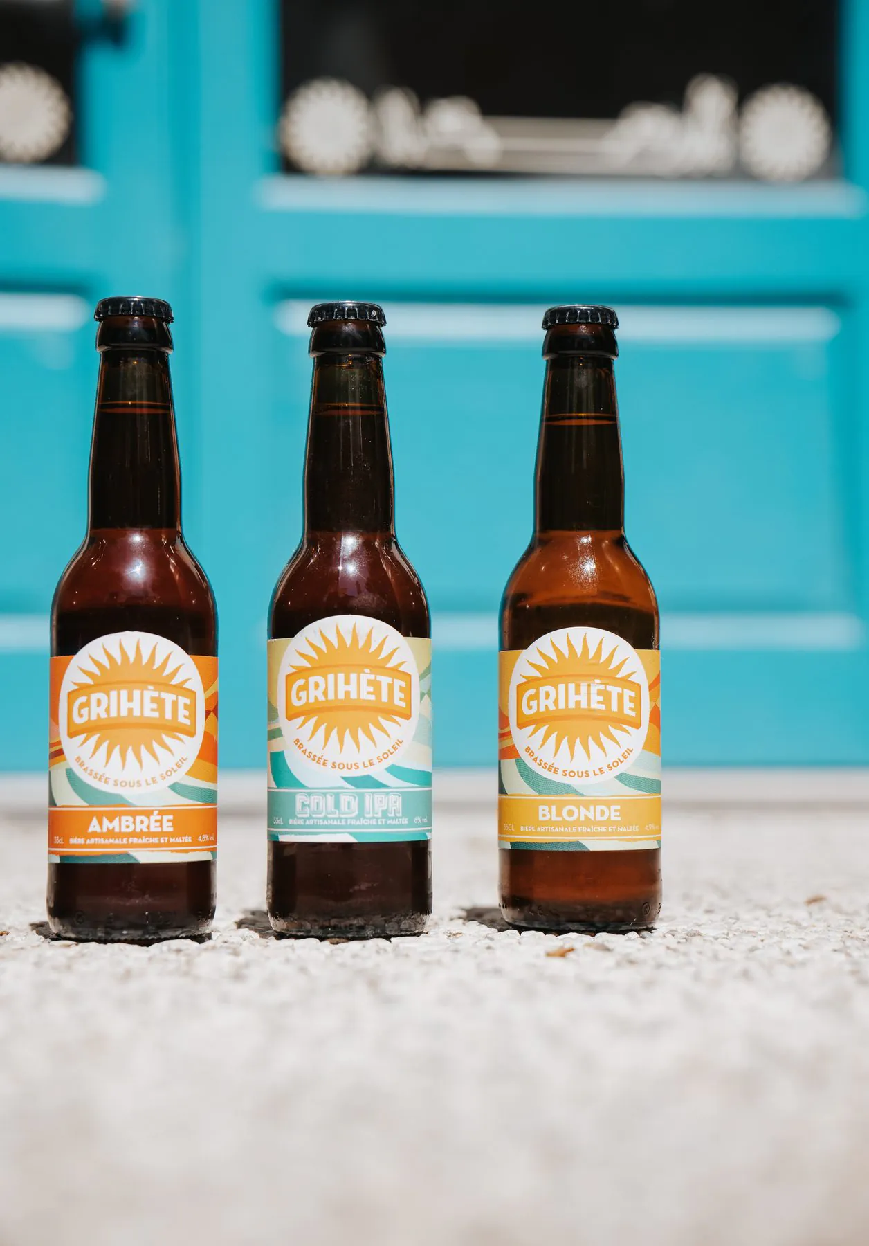 Trois bières artisanales Grihète, ambrée, cold IPA et blonde, devant la porte turquoise