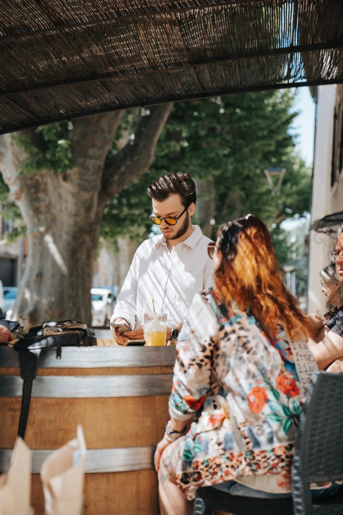 Léo Ricard derrière le tonneau du bar, sous les platanes, avec des clients en terrasse