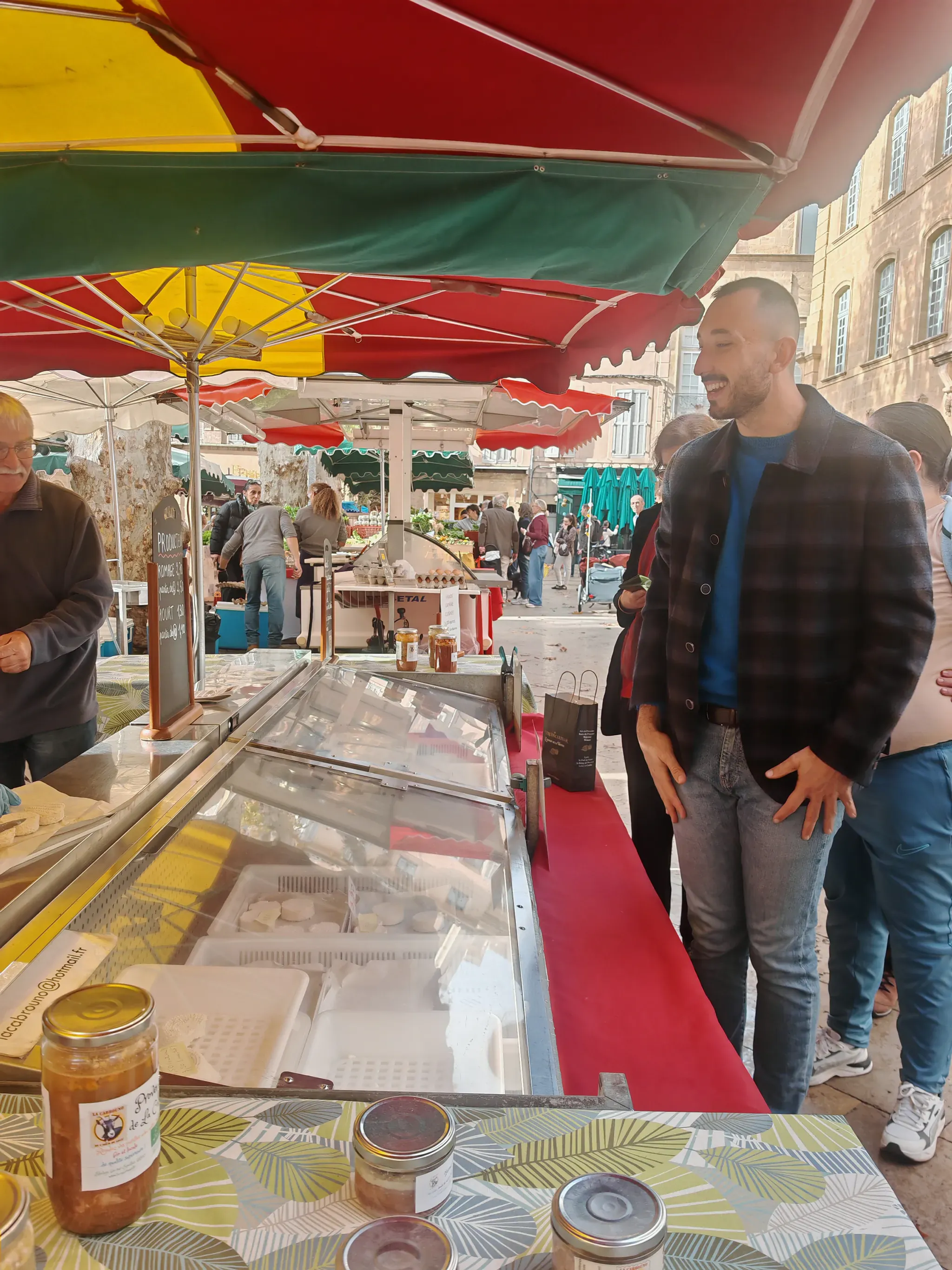 Commercant souriant devant son etal de fromages et de conserves sur un marche provencal d'Aix