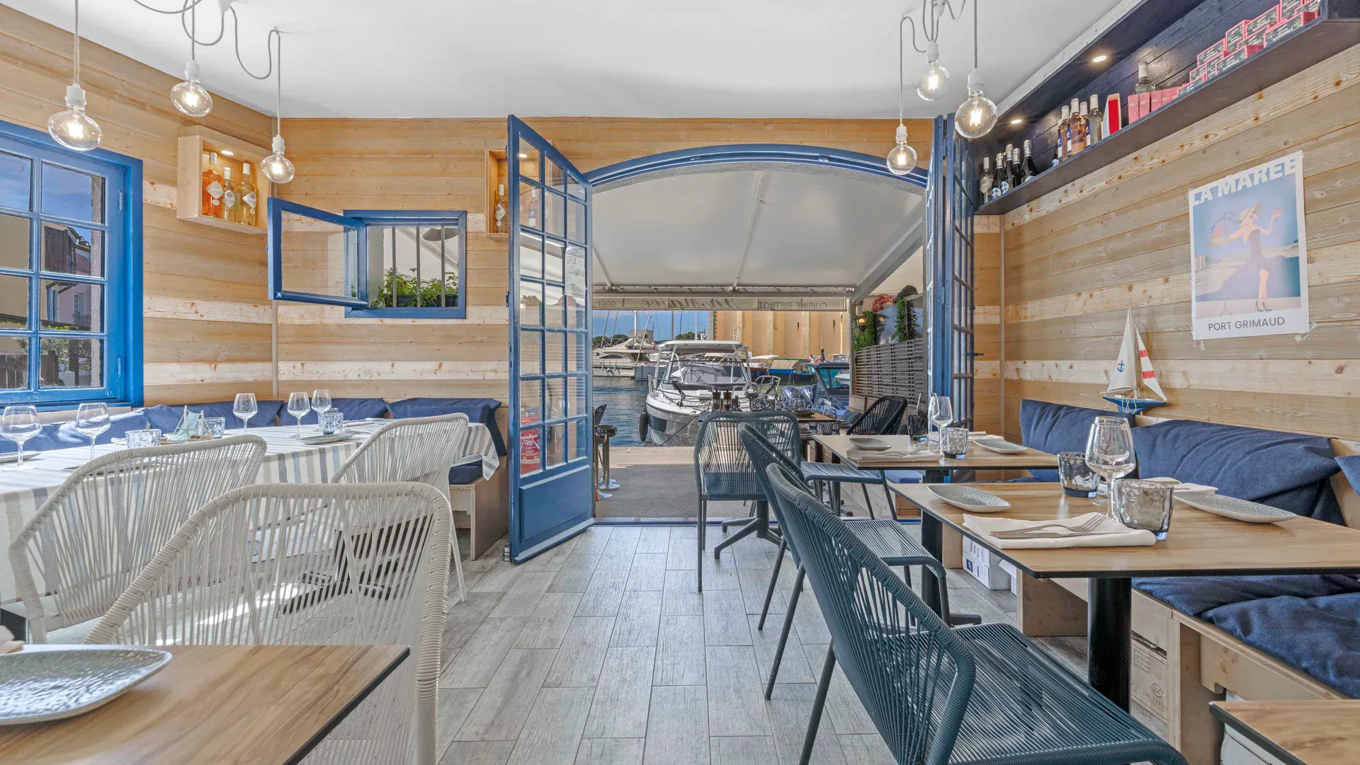 Salle intérieure du restaurant La Marée à Port Grimaud, bois clair, banquettes bleu marine et vue sur les bateaux du port