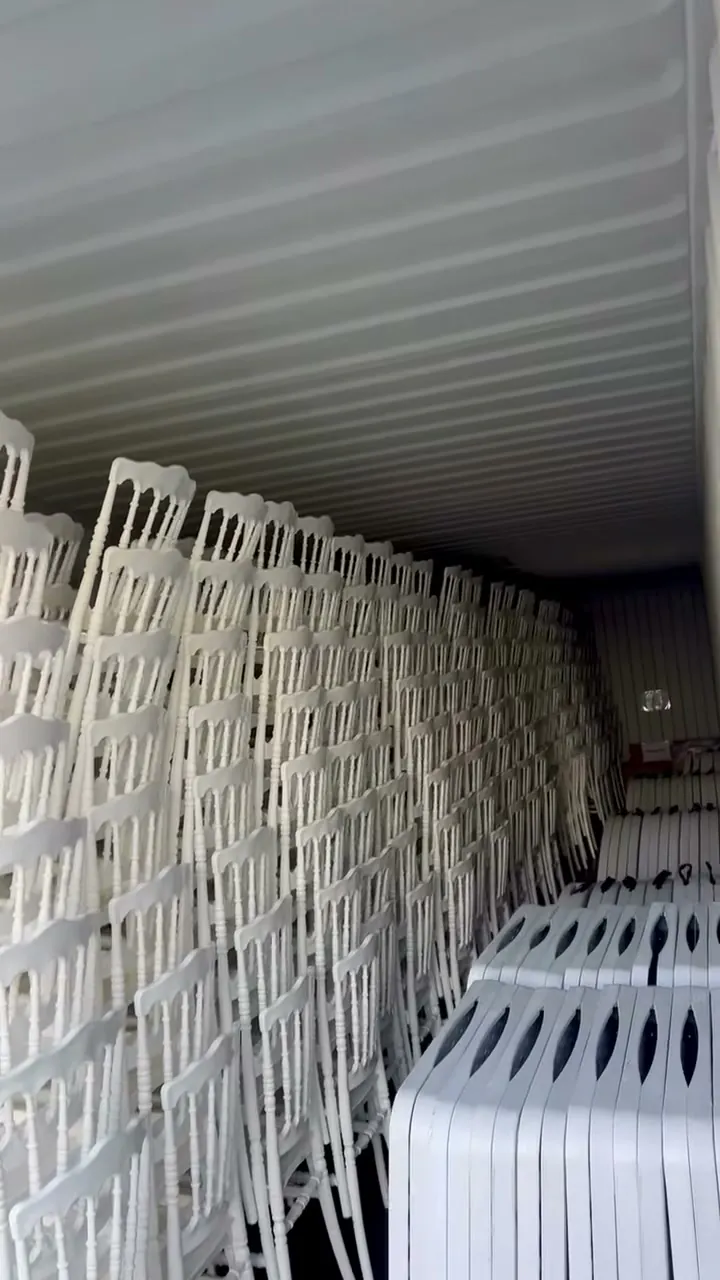 Intérieur du dépôt : longue enfilade de chaises Napoléon blanches empilées le long de la paroi, tables pliantes rangées au sol.