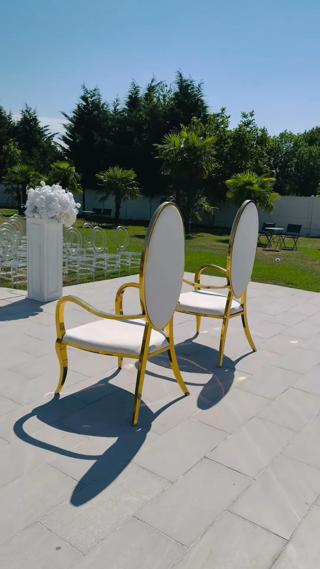 Deux fauteuils médaillon à structure dorée et assise blanche posés sur une terrasse dallée, à côté d'une colonne blanche surmontée de fleurs blanches.