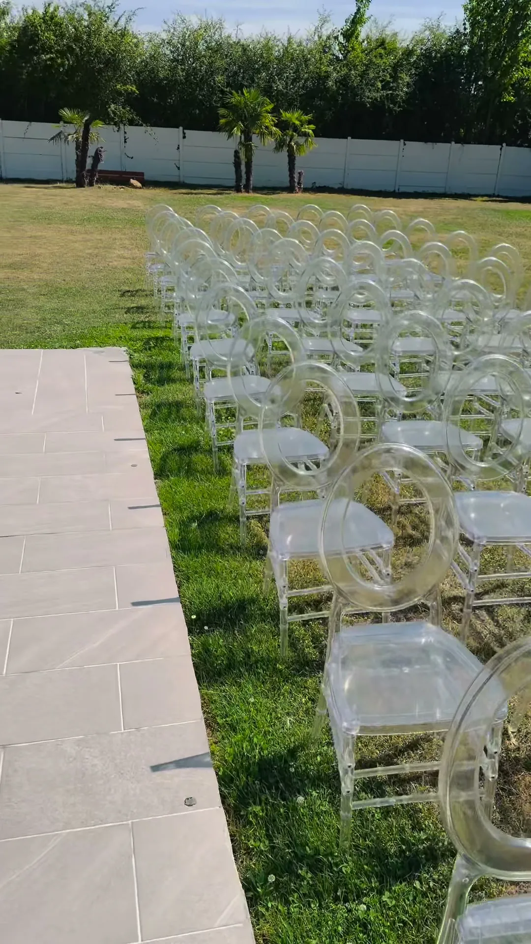 Rangées de chaises transparentes à dossier médaillon alignées sur une pelouse, de part et d'autre d'une allée dallée blanche.