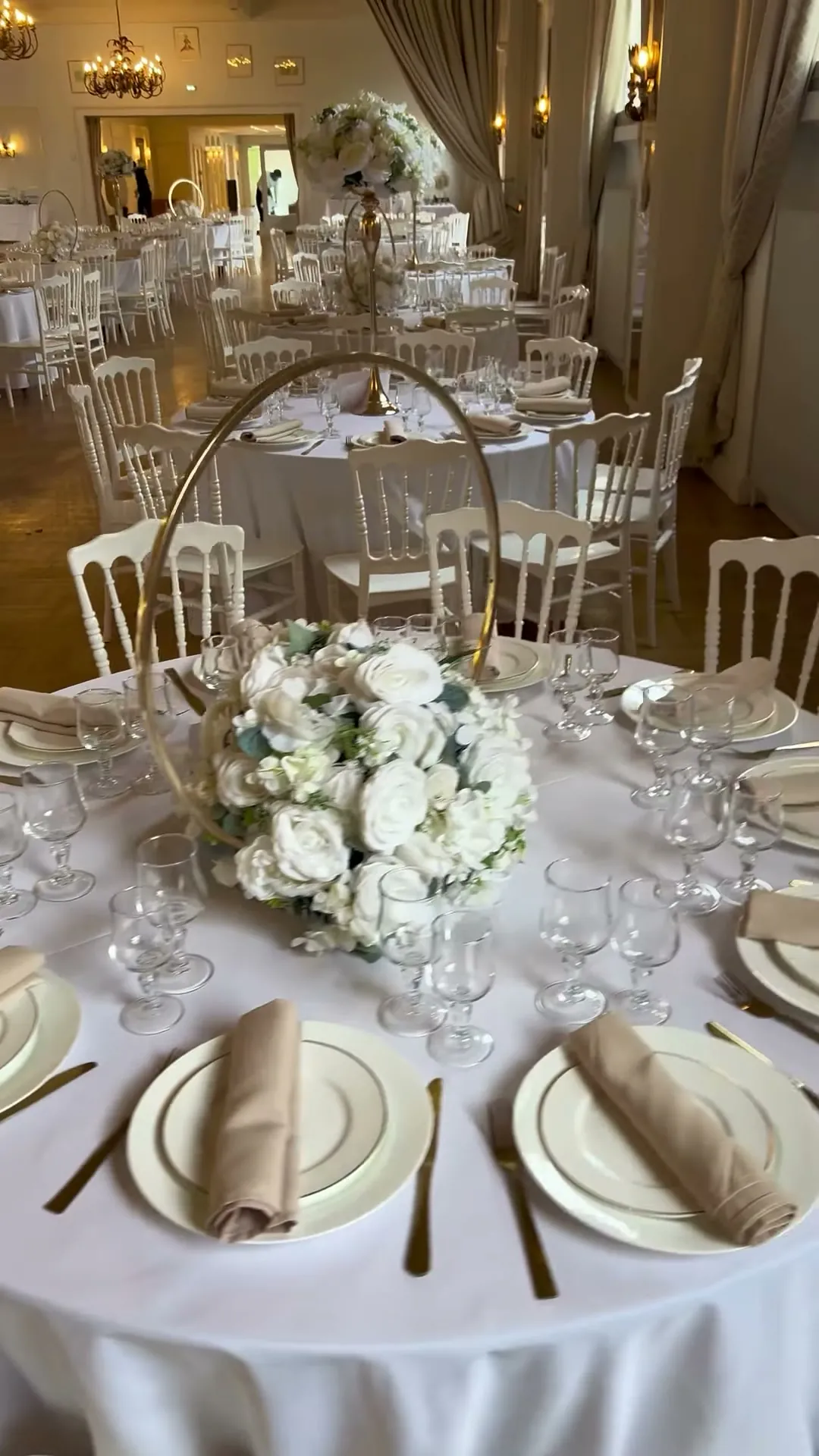 Détail de dressage : assiettes empilées, serviettes beiges roulées, couverts dorés, verrerie et centre de table de roses blanches monté sur un arceau doré.