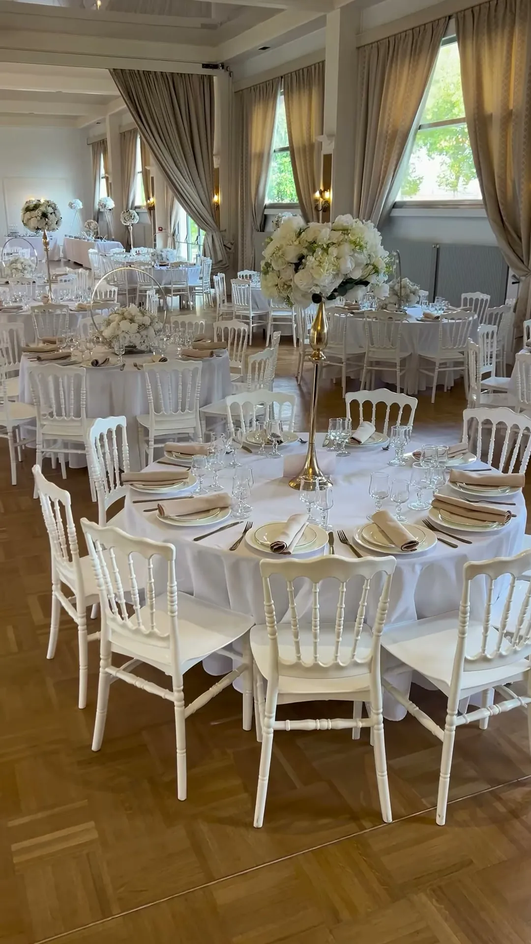 Table ronde nappée de blanc entourée de chaises Napoléon, candélabre doré surmonté d’un bouquet blanc, assiettes, serviettes beiges et verrerie.