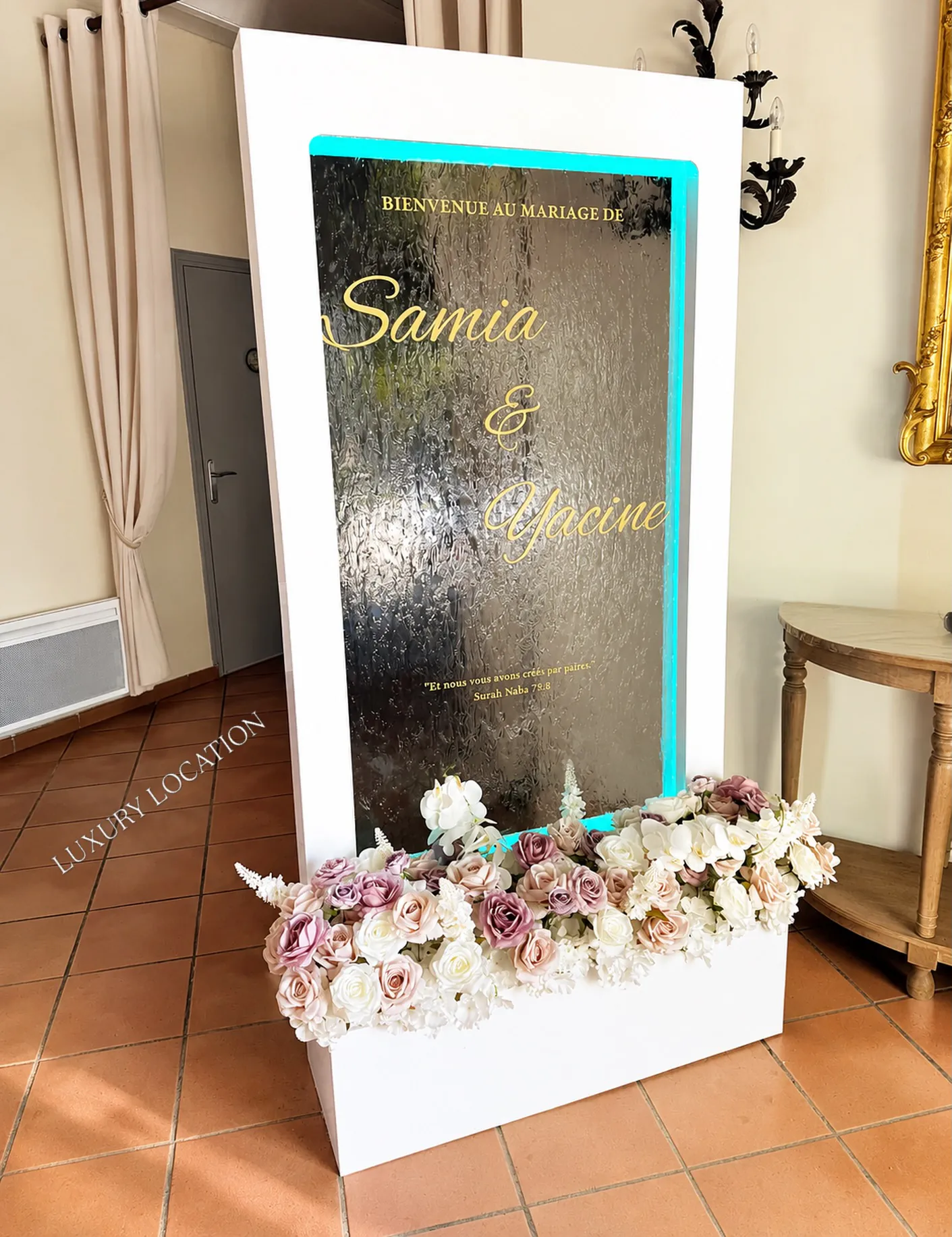 Miroir de bienvenue personnalisé au prénom des mariés, bordé d'un ruban lumineux et d'une jardinière de fleurs, installé à l'entrée d'une salle de réception.