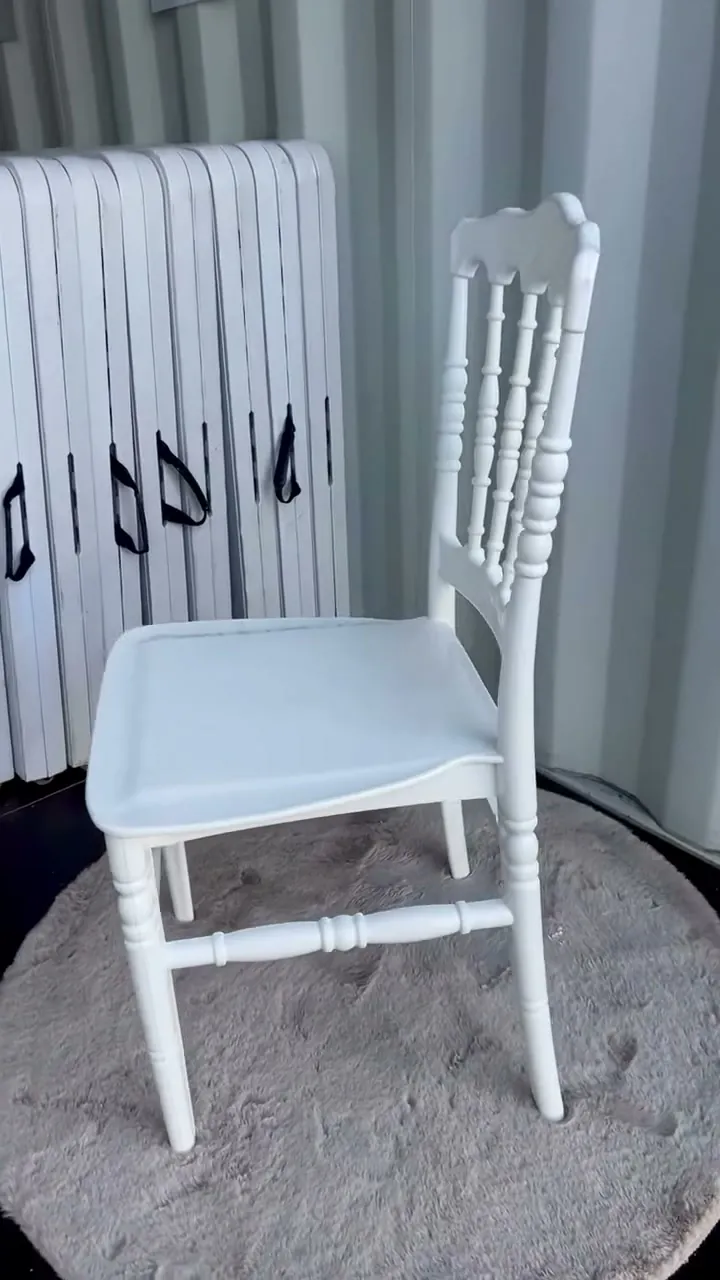 Une chaise Napoléon blanche vue de trois quarts, posée sur un tapis rond, devant des tables pliantes rangées au dépôt.
