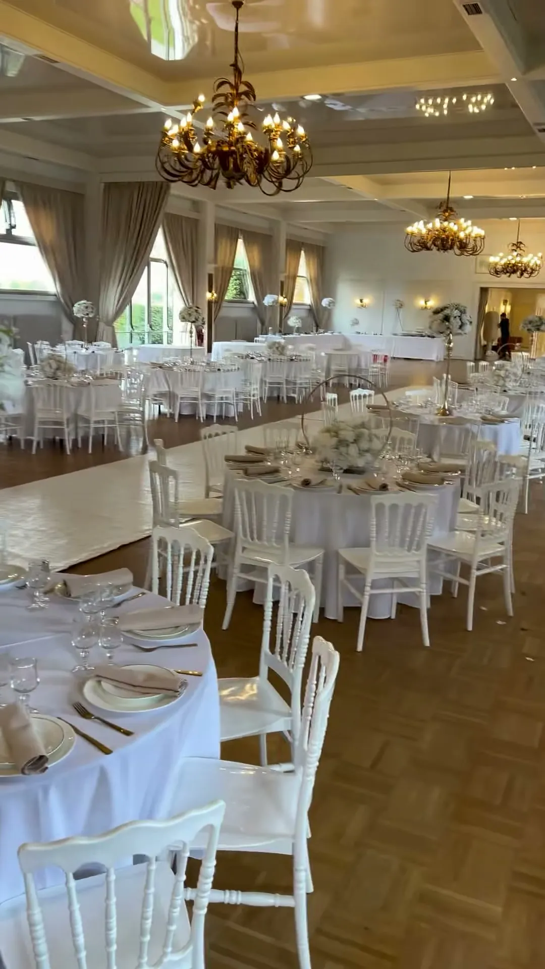 Vue d'ensemble d'une salle de réception : tables rondes nappées de blanc, table d'honneur rectangulaire, chaises Napoléon, lustres dorés et hautes fenêtres.