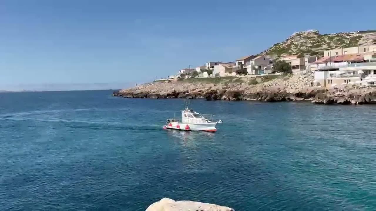 Bateau de pêche naviguant près d'une côte rocheuse méditerranéenne avec un village en arrière-plan