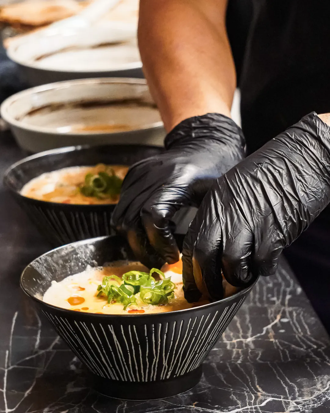 Mains gantées dressant un bol de ramen sur un plan de travail en marbre