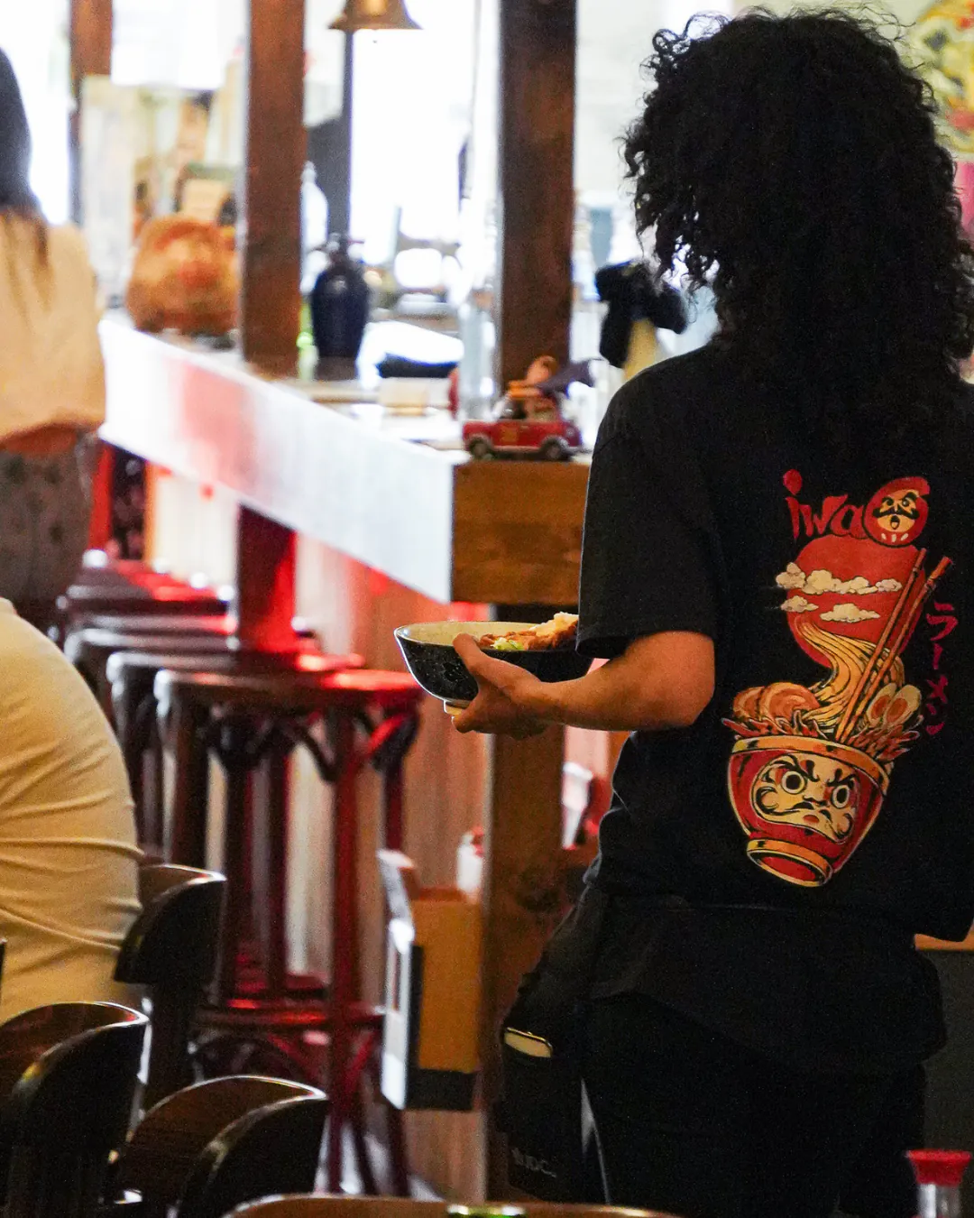 Serveuse d'Iwao de dos, t-shirt daruma-ramen, portant un bol vers le comptoir