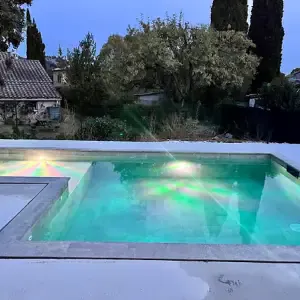 Piscine résidentielle éclairée en soirée devant une maison provençale et son jardin arboré