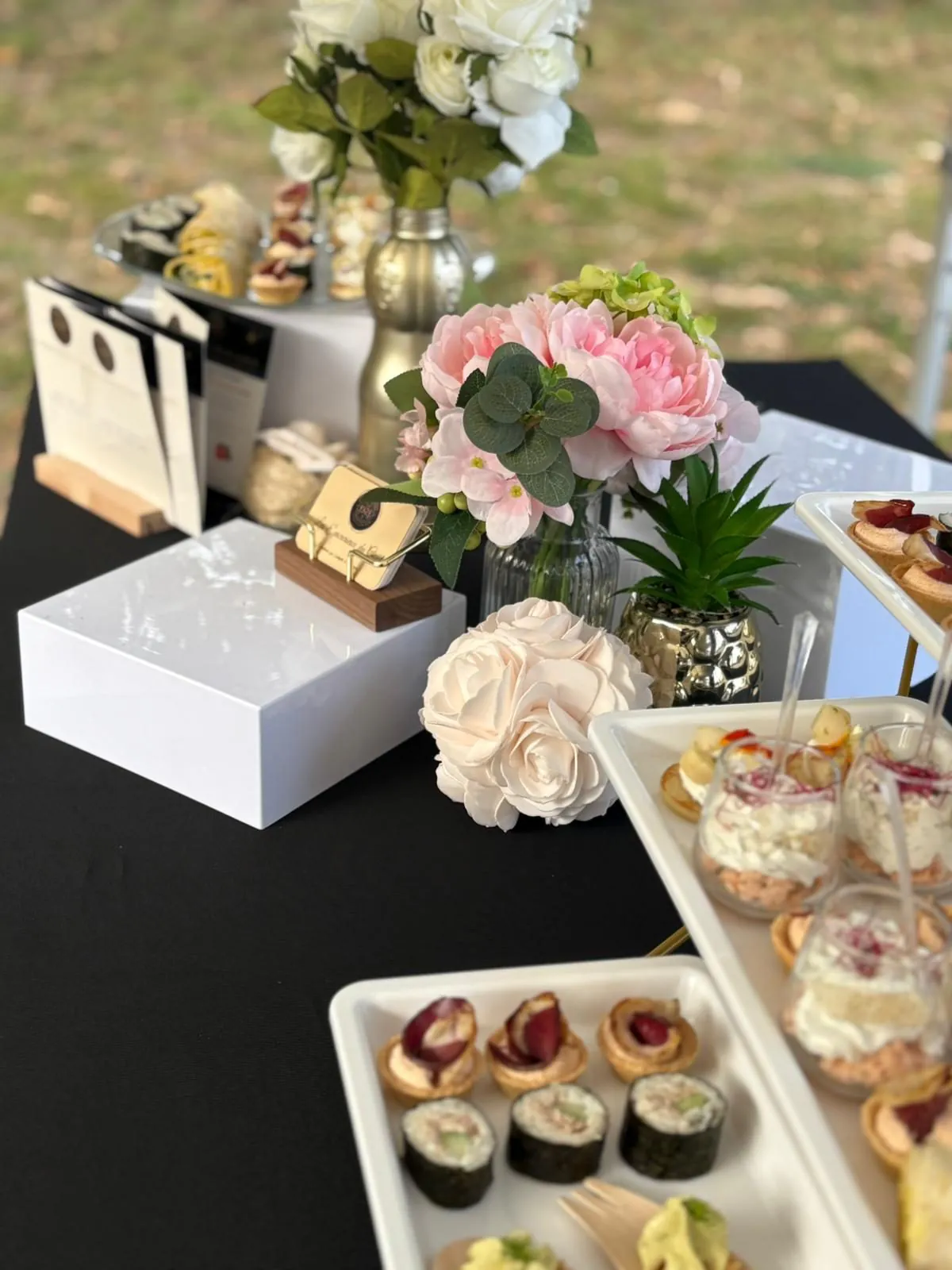 Table traiteur en extérieur sur nappe noire, plateaux de makis, tartelettes et bouchées décorés de fleurs