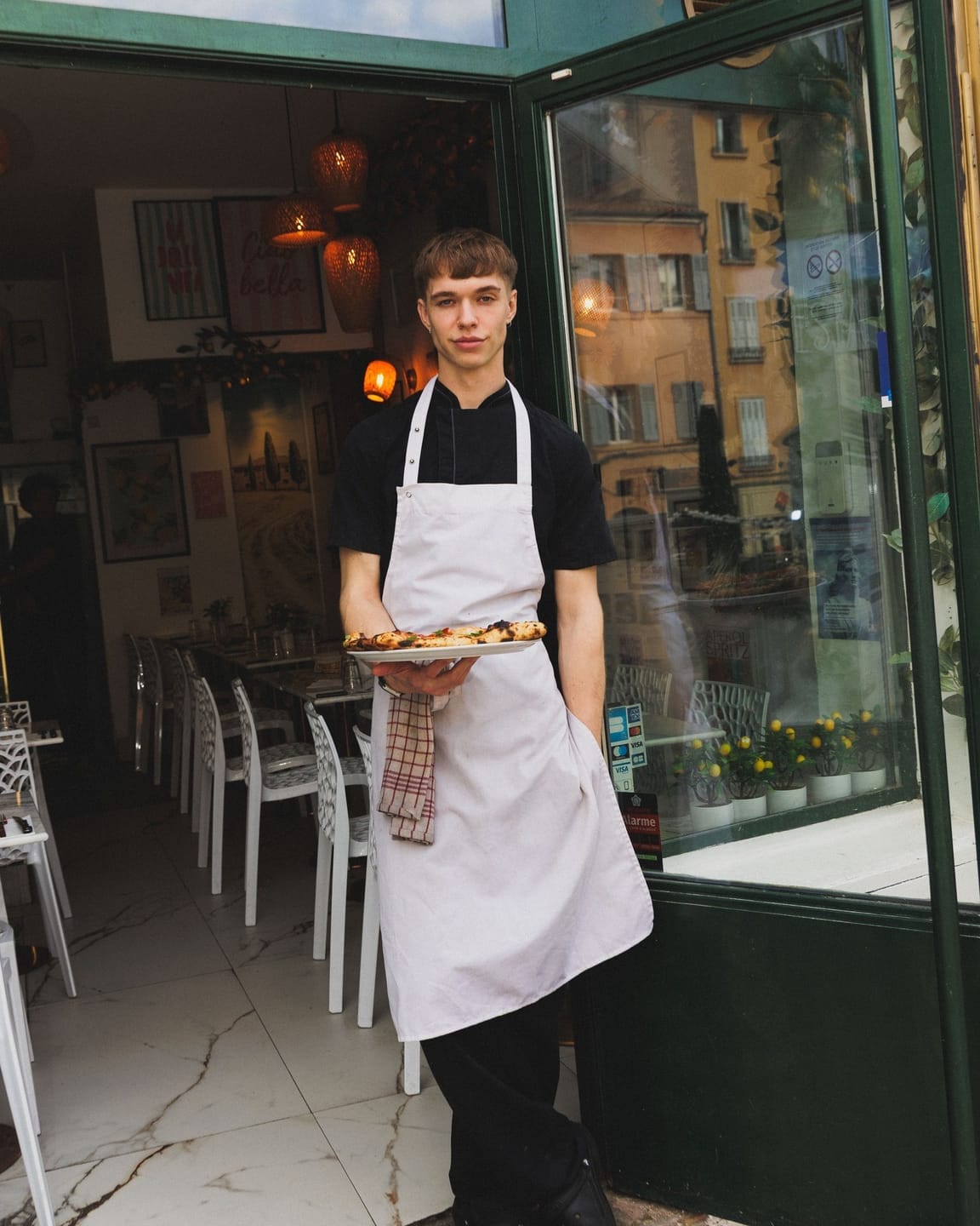 Jeune serveur en tablier blanc présentant une assiette de pizza à l'entrée du restaurant italien Di Micheli